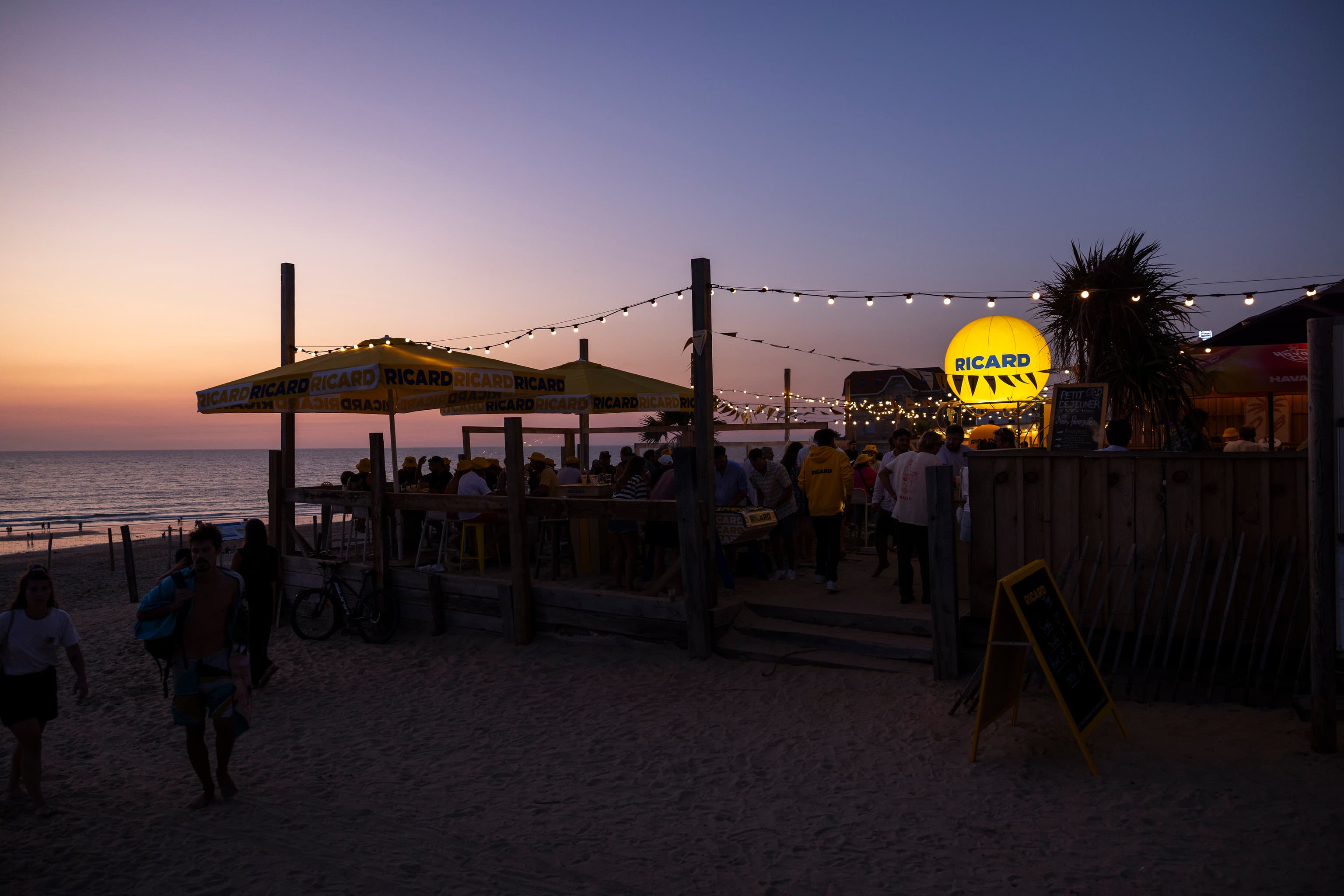 Bar de plage habillé aux couleurs Ricard au coucher de soleil, parasols et ballon lumineux, lors du Ricard Summer Tour 2026