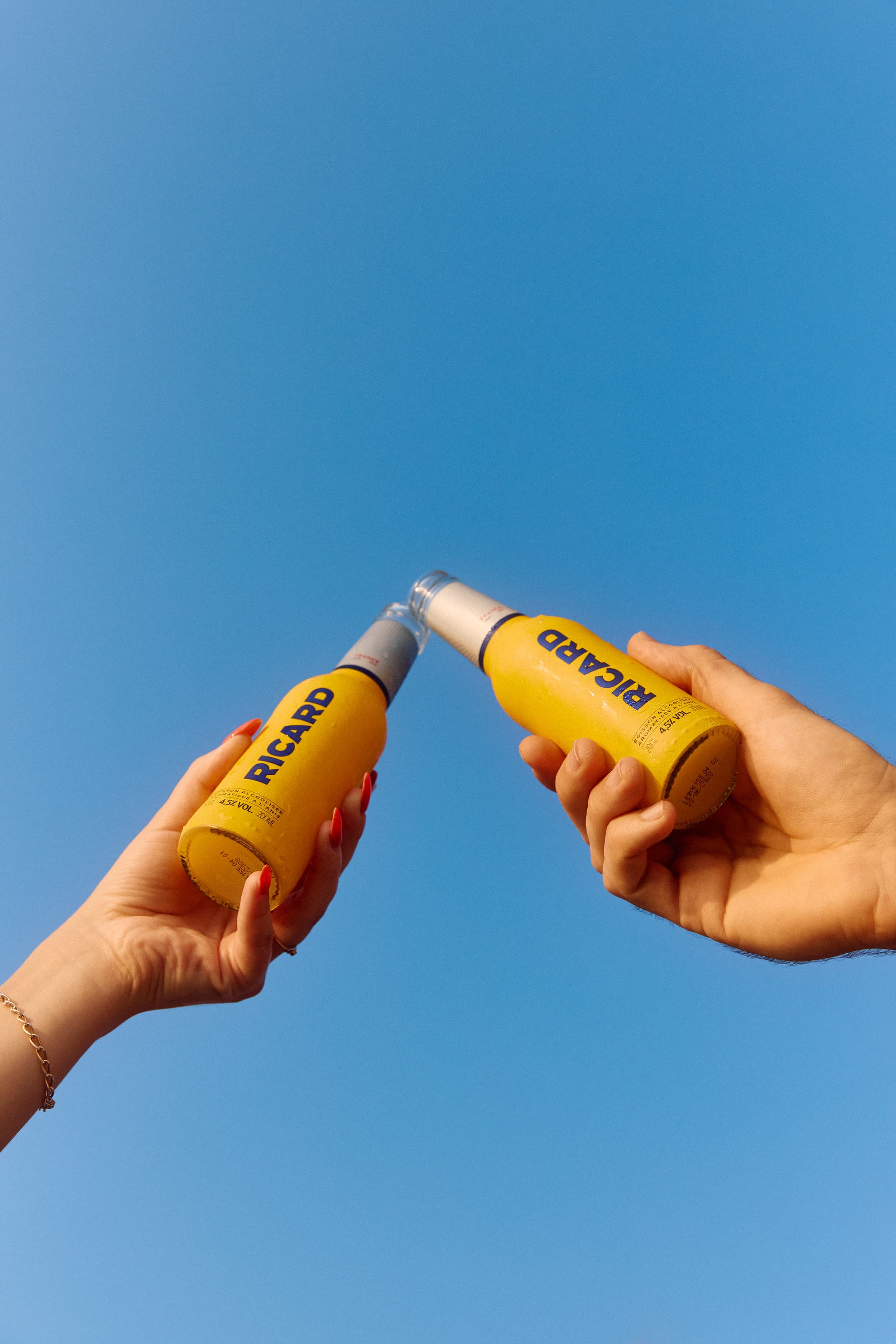 Deux mains trinquant avec des bouteilles Ricard sur fond de ciel bleu pendant le Ricard Summer Tour 2026