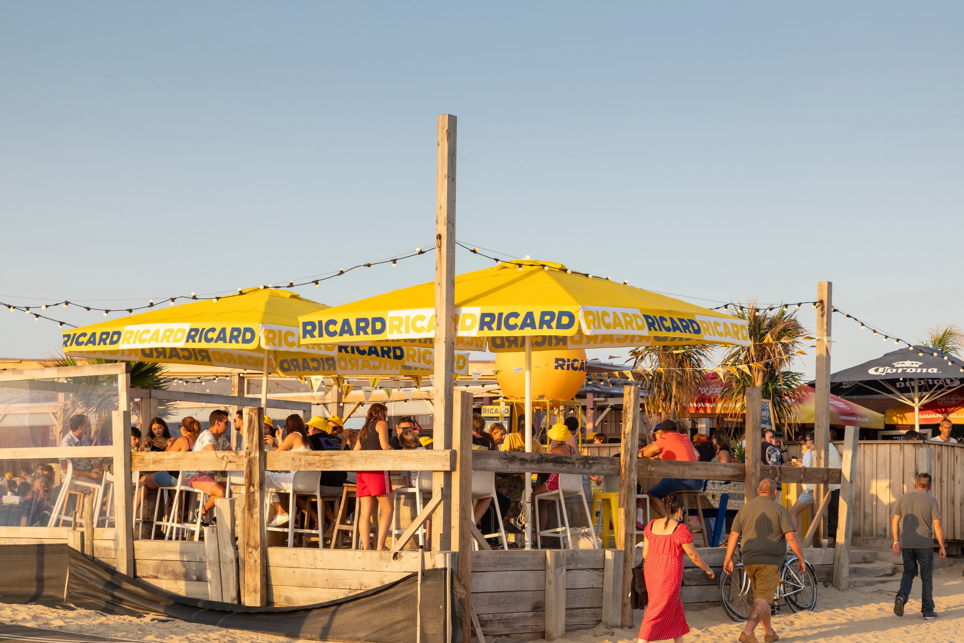 Bar de plage Ricard en pleine journée avec parasols jaunes et clients attablés, Ricard Summer Tour 2026
